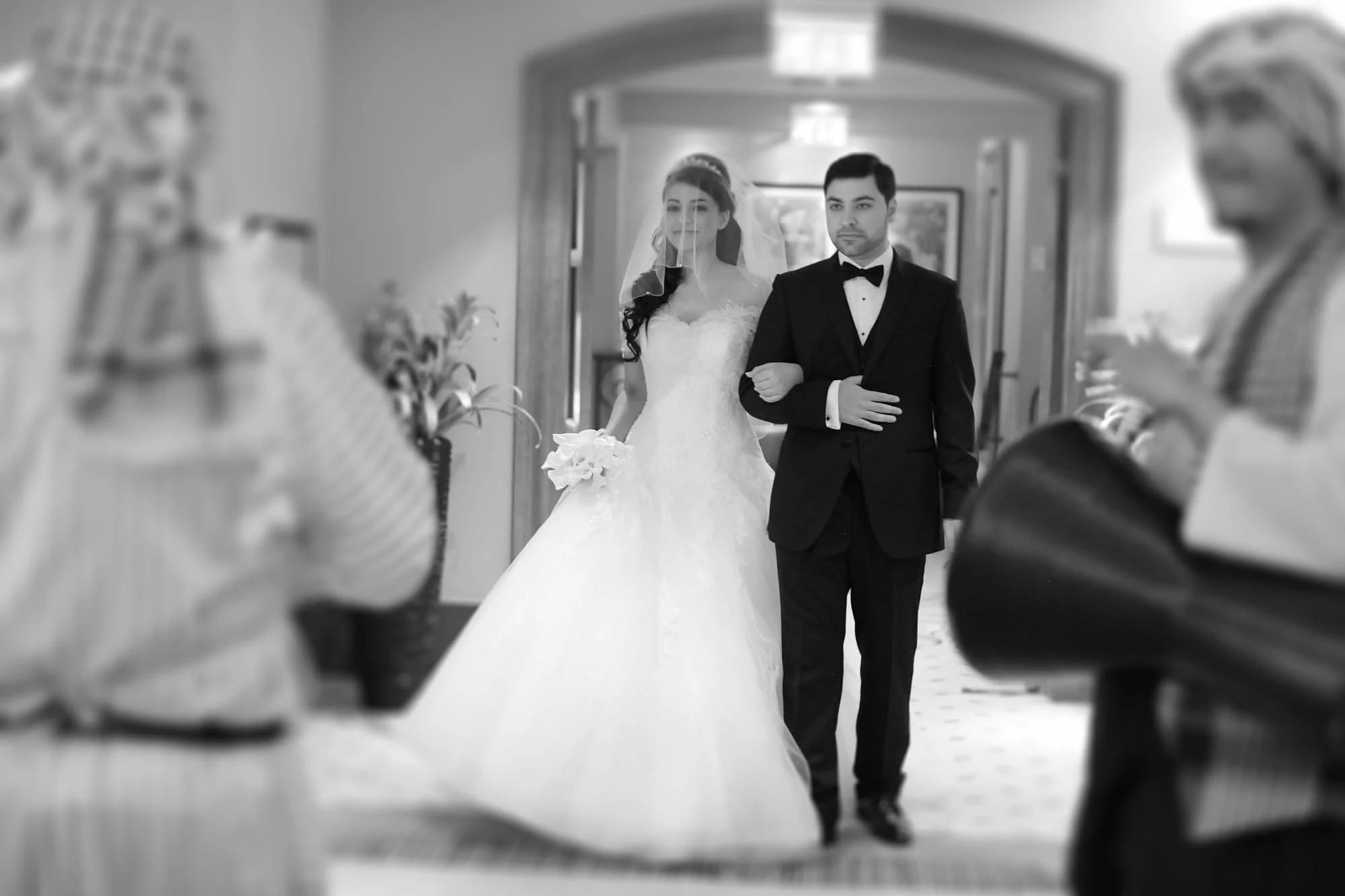 A bride in a full white gown walks arm in arm with a man in a tuxedo along a hotel corridor past blurred guests.