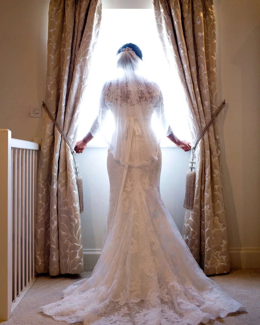 A bride in a lace gown with her back to the camera, holding the curtains of a bright window