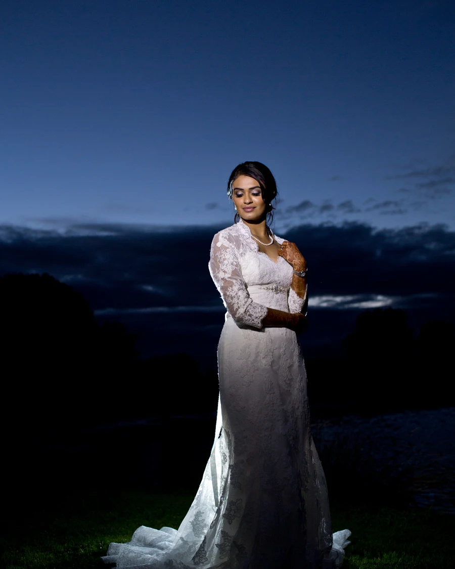 A bride in a long-sleeved lace gown poses on a lawn under a deep blue twilight sky.