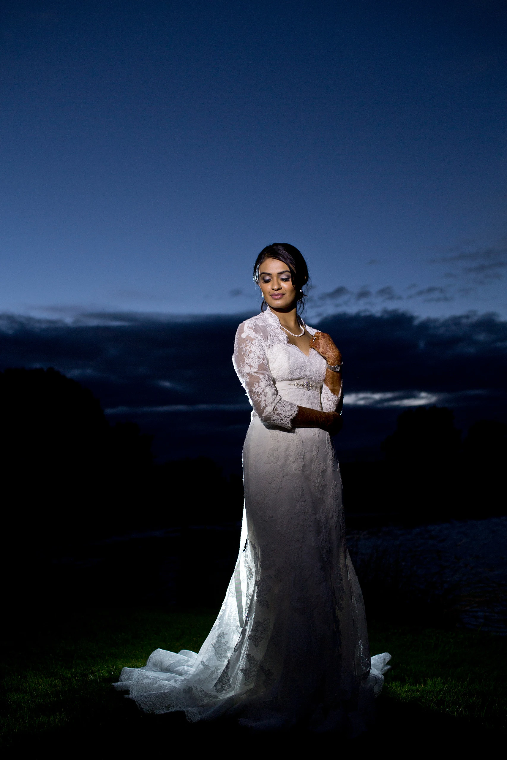 A bride in a long-sleeved lace gown poses on a lawn under a deep blue twilight sky.