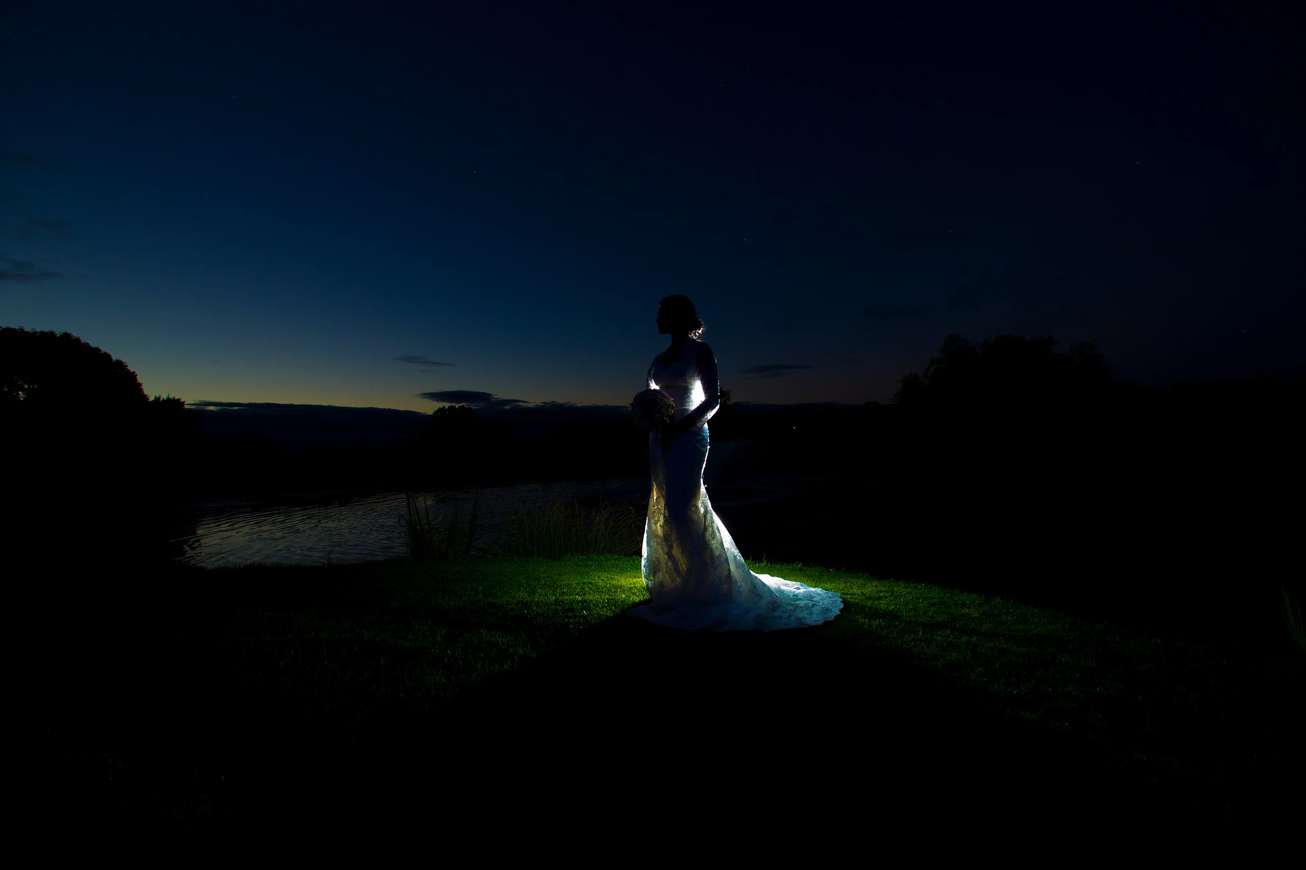 A bride in a white gown is lit against a dark blue night sky and treeline beside water.