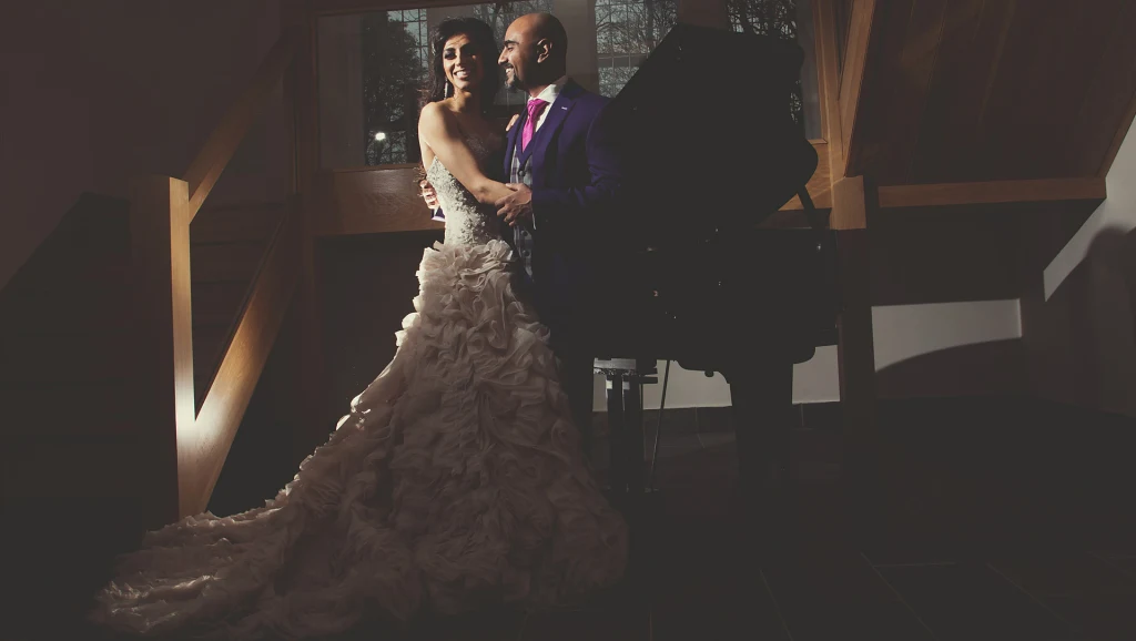 A bride in a ruffled gown and her groom embrace beside a grand piano