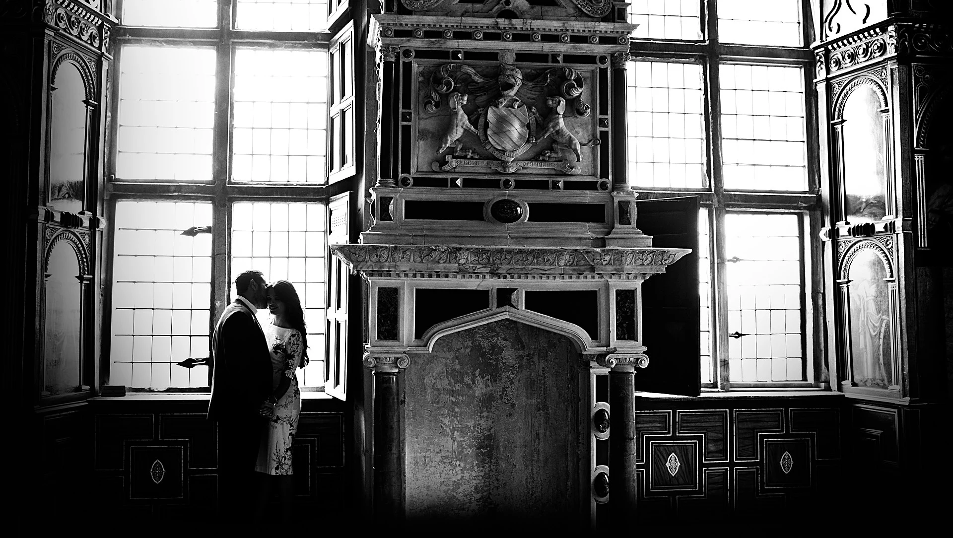 A couple kiss in silhouette at a tall leaded window beside a carved fireplace in a historic panelled room.