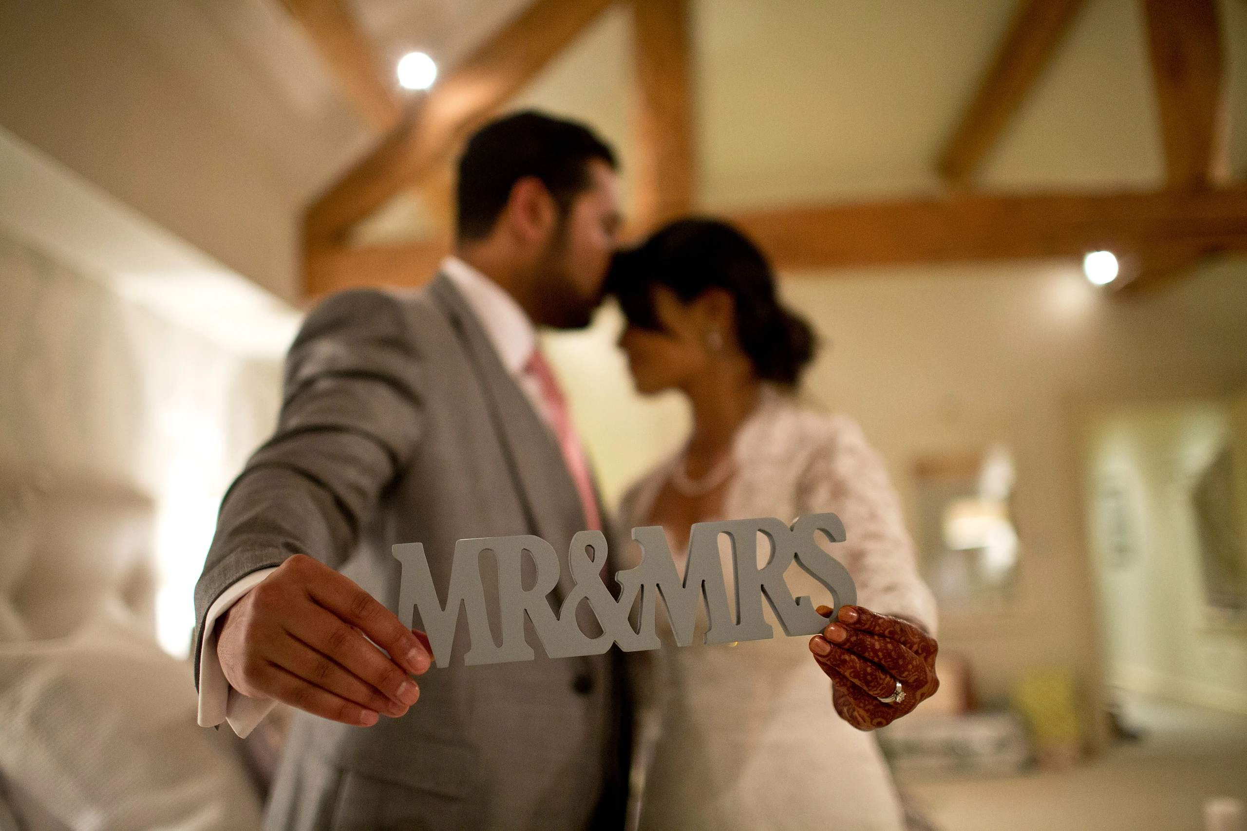 A groom in a grey suit presses his forehead to his bride's while they hold a silver Mr and Mrs sign, in a beamed room.