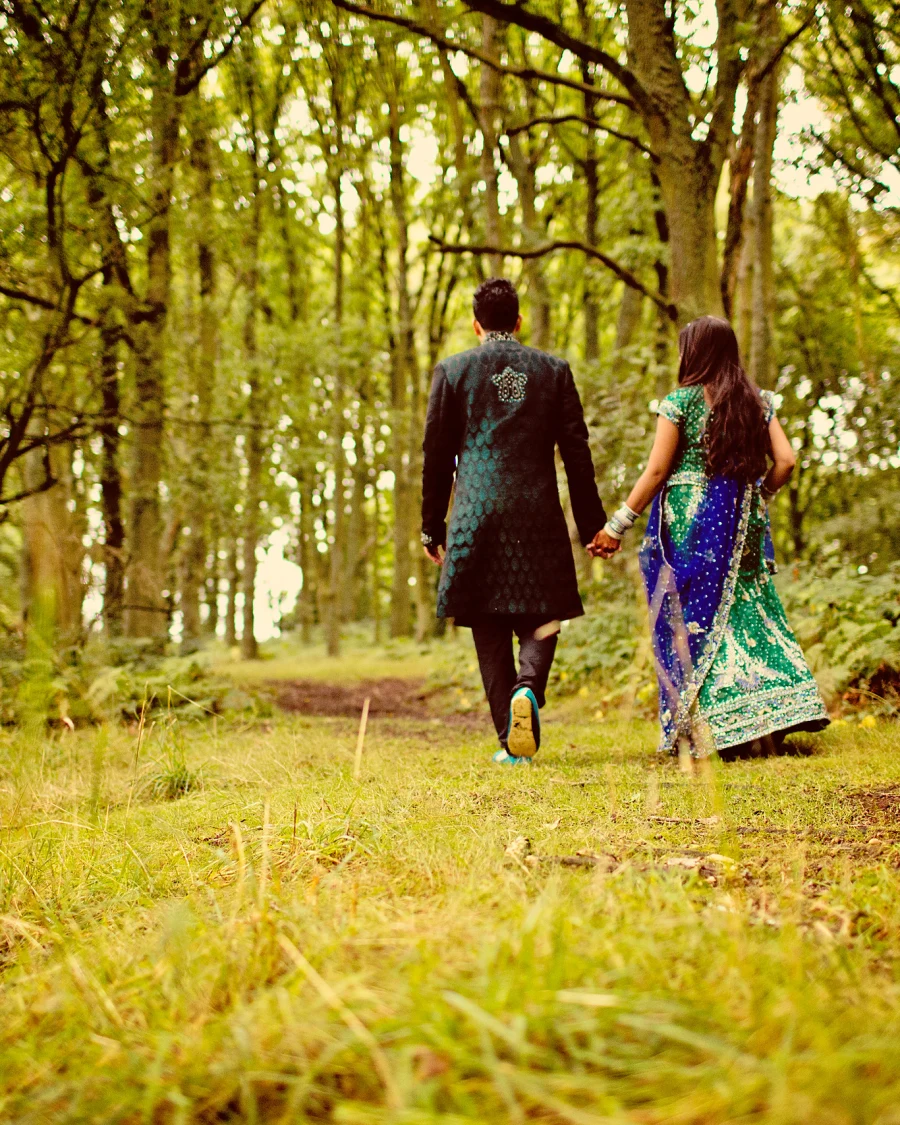 A couple in Asian wedding clothes walk away hand in hand along a grassy woodland path.