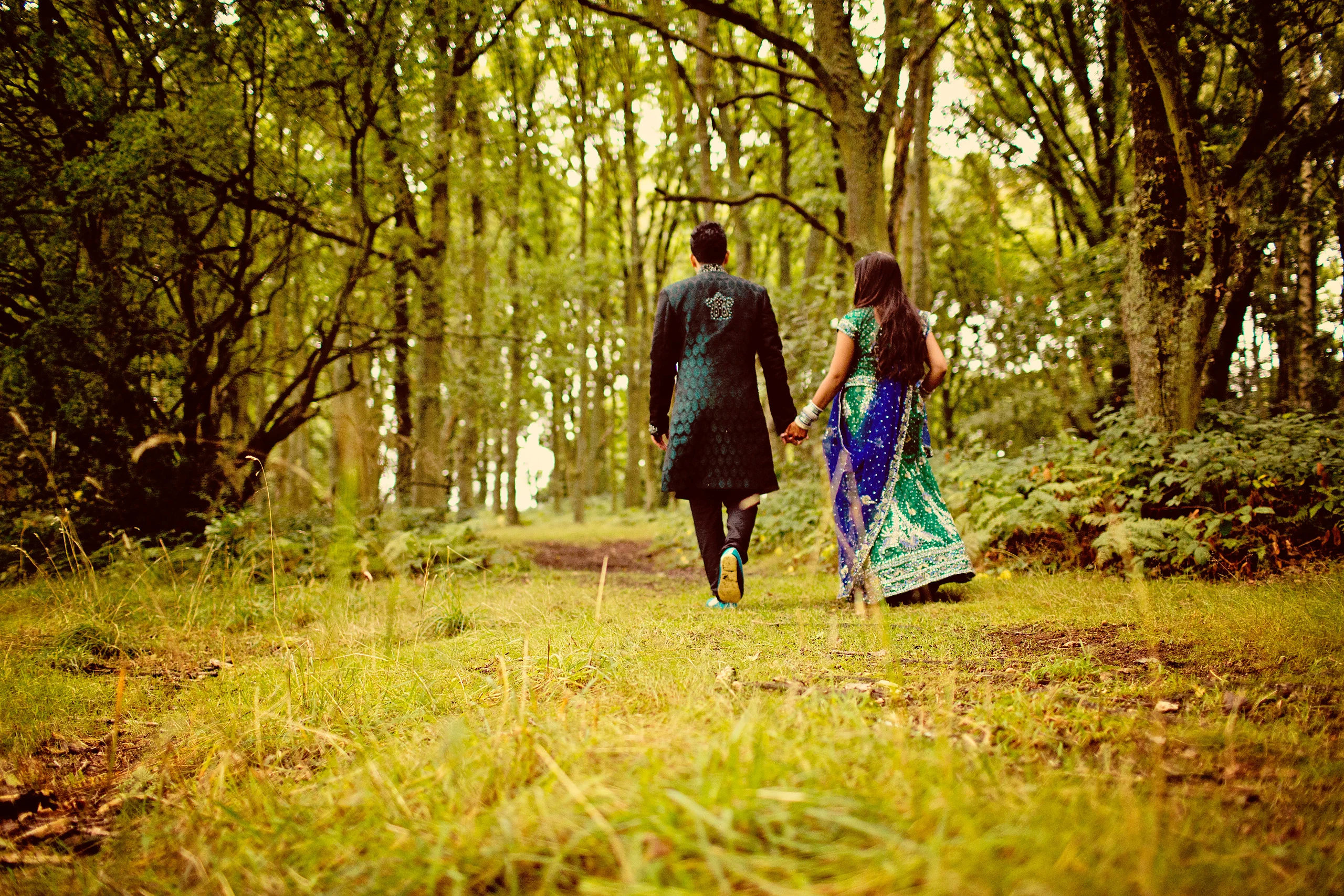 A couple in Asian wedding clothes walk away hand in hand along a grassy woodland path.