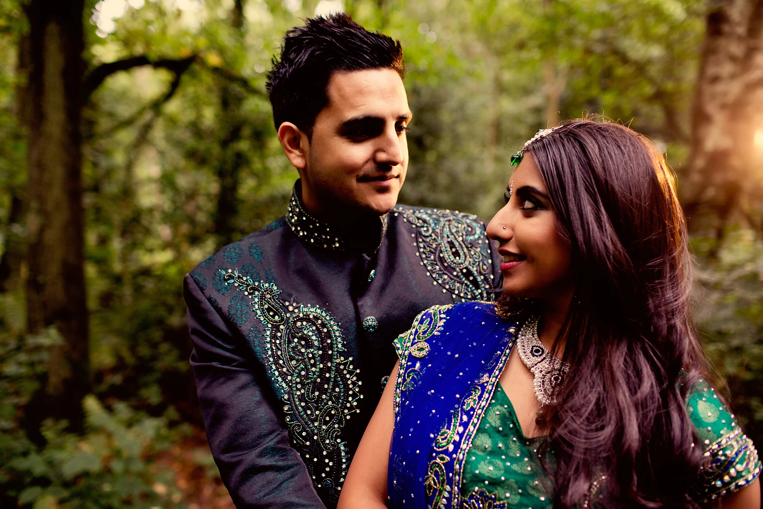 A smiling groom in an embroidered sherwani holds his bride in a blue sari among woodland ferns.