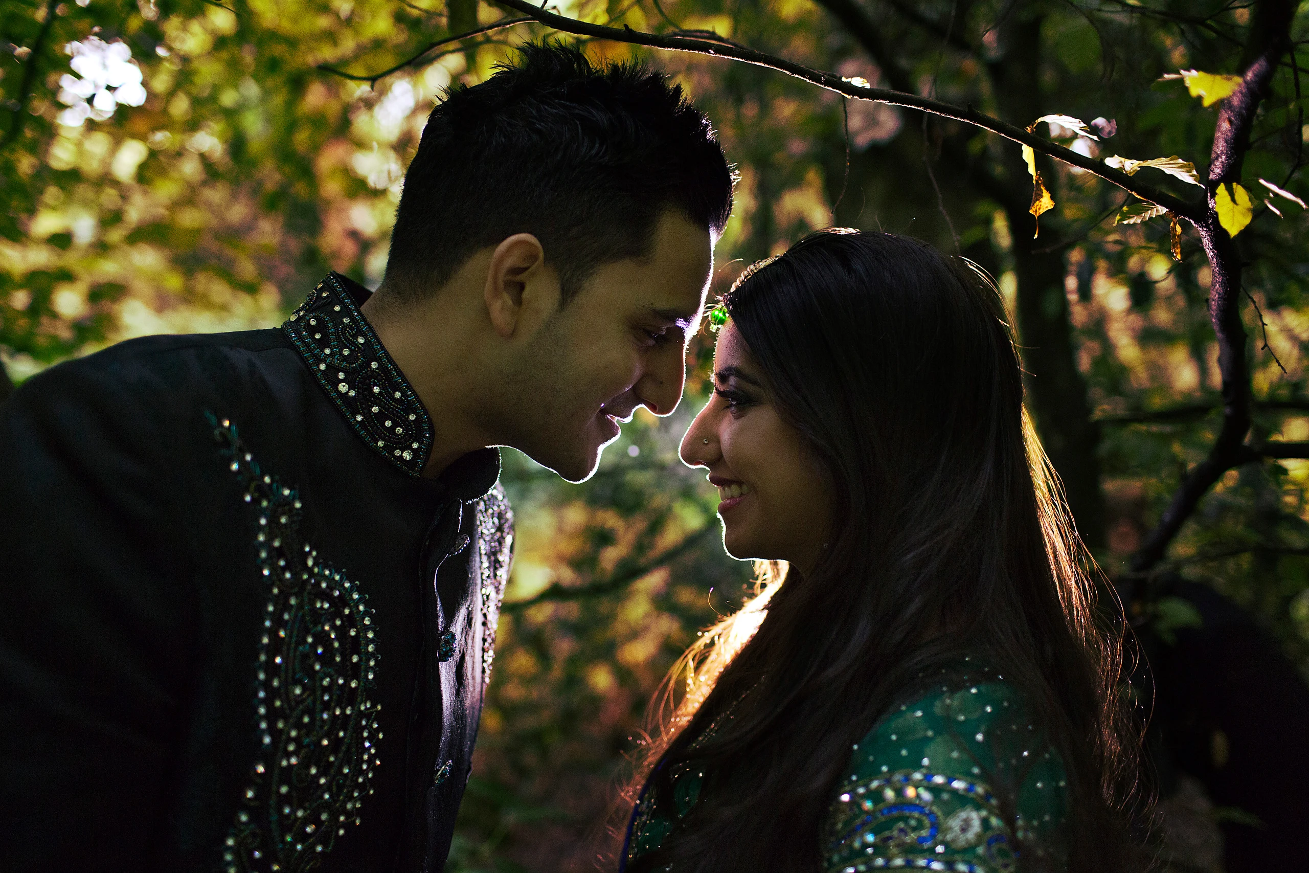 A groom in an embellished sherwani and a bride with long hair stand nose to nose under woodland branches.