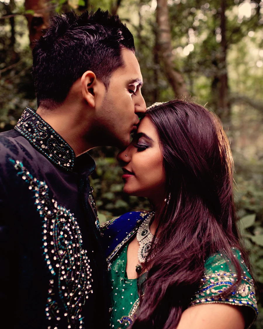 In woodland a groom kisses the forehead of his bride in a green and blue sari