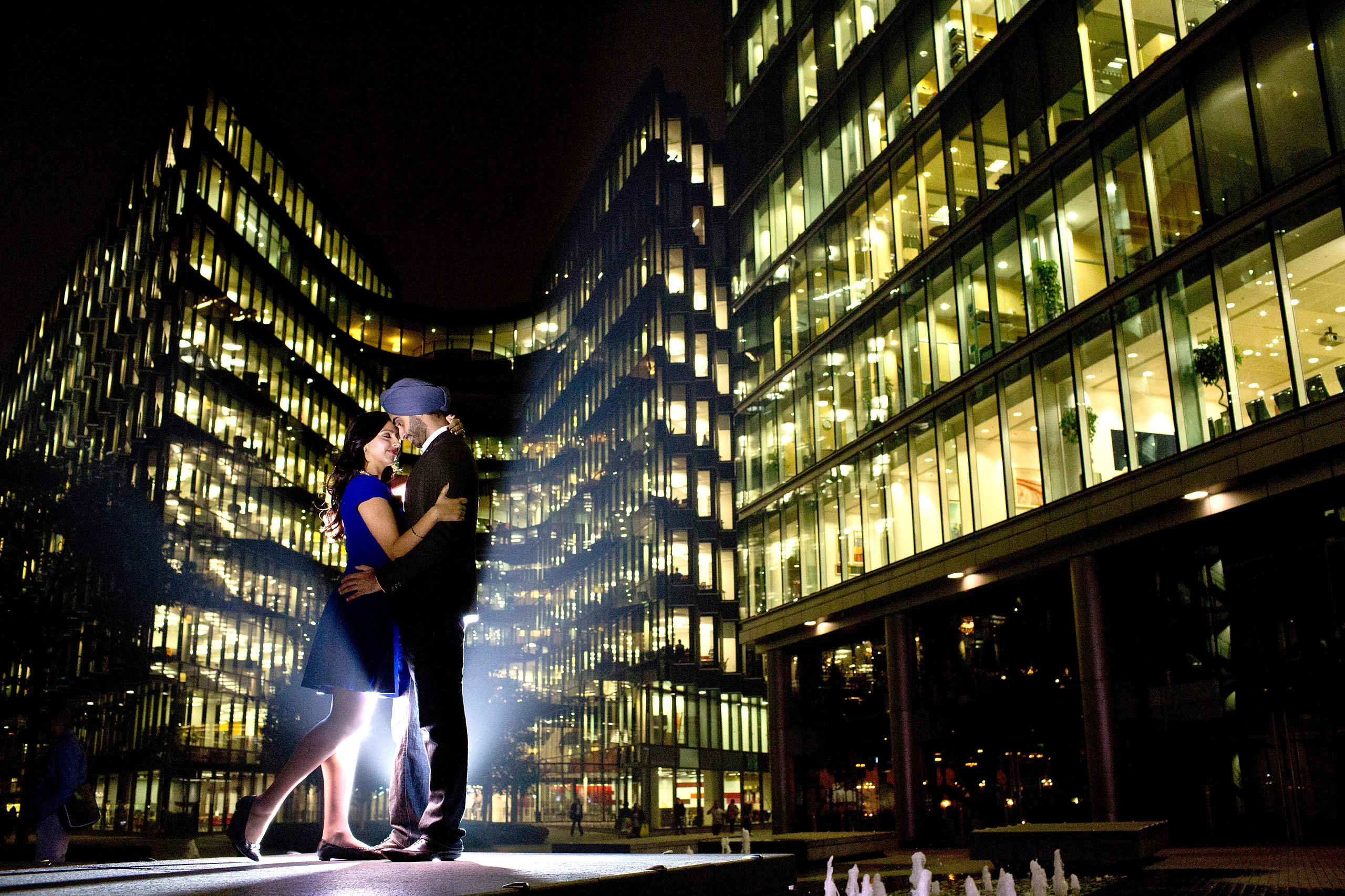A couple embrace in a pool of light between glowing glass office buildings in the City at night.