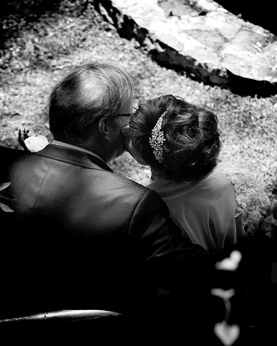 An older bride and groom share a kiss on a garden bench, seen from behind in dramatic black and white.