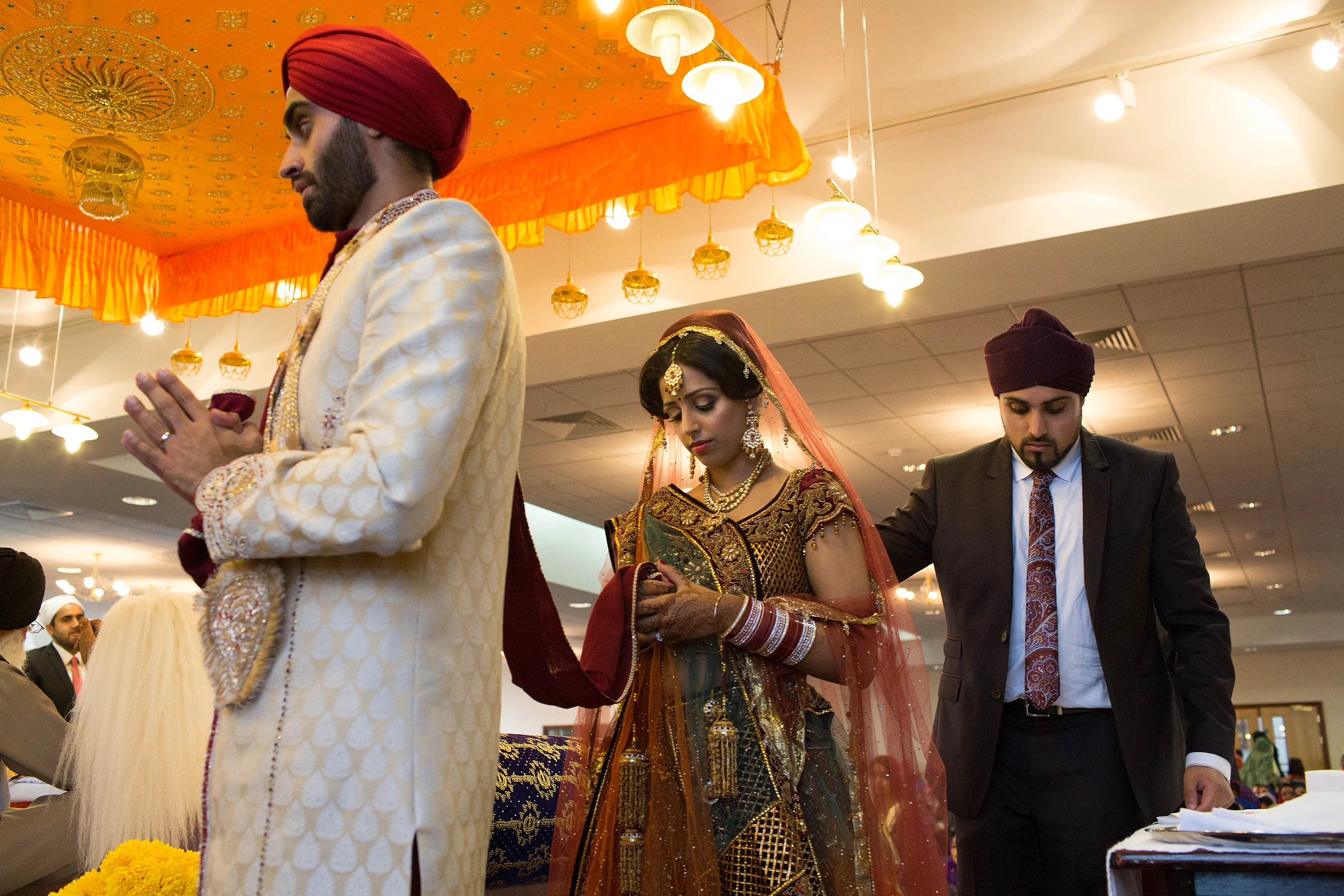 A Sikh groom in a cream sherwani and red turban stands with palms together beside his bride in a red and gold lehenga during the ceremony.