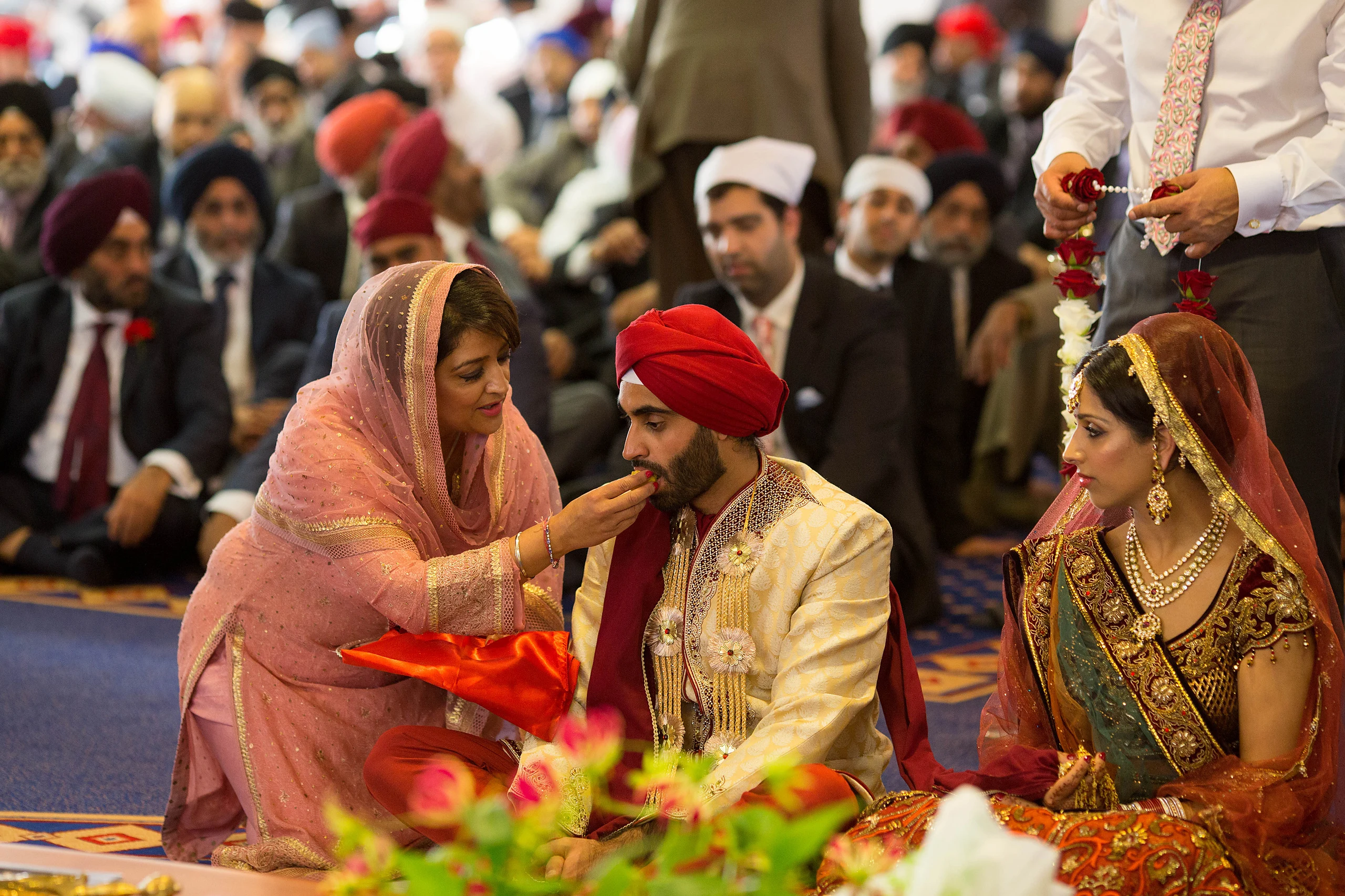 During a Sikh ceremony a woman in a pink suit feeds the groom while the bride sits beside him and turbaned guests look on.