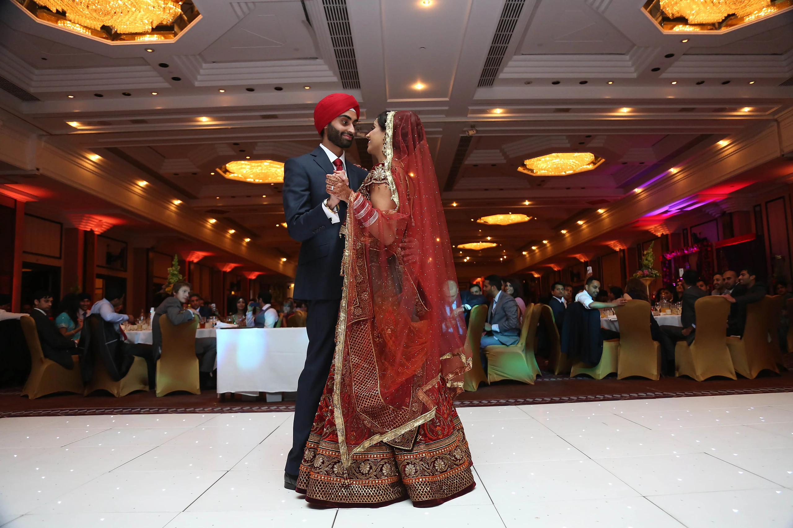 A turbaned groom and his bride in a red lehenga share a first dance on a white dance floor in a large banqueting hall.