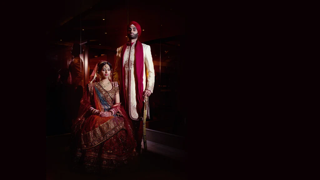 A seated bride in a red lehenga and her turbaned groom in a dark, moody portrait