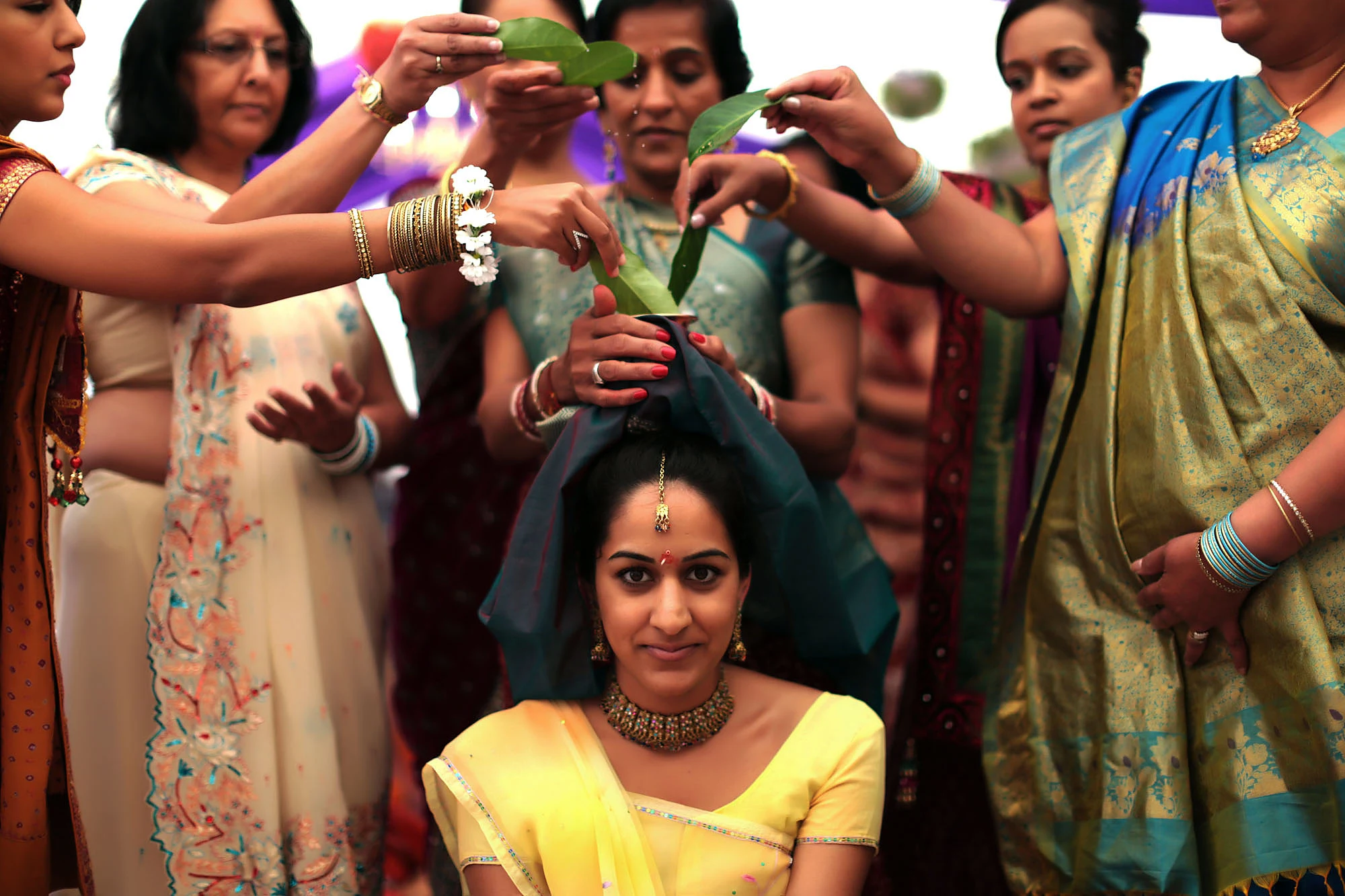 Women in saris hold leaves over the head of a seated bride in yellow during a pre-wedding blessing.