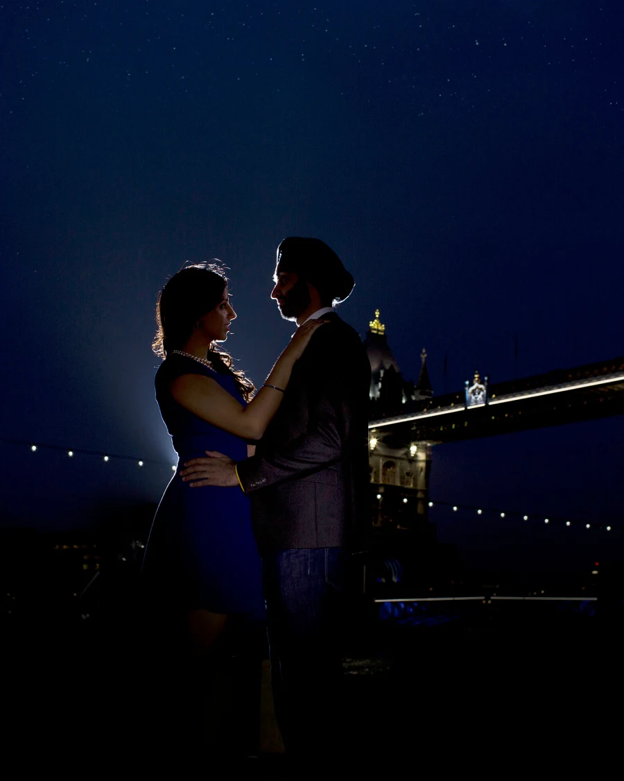A couple embrace on the Thames riverside at night with Tower Bridge lit behind them under a starry blue sky.