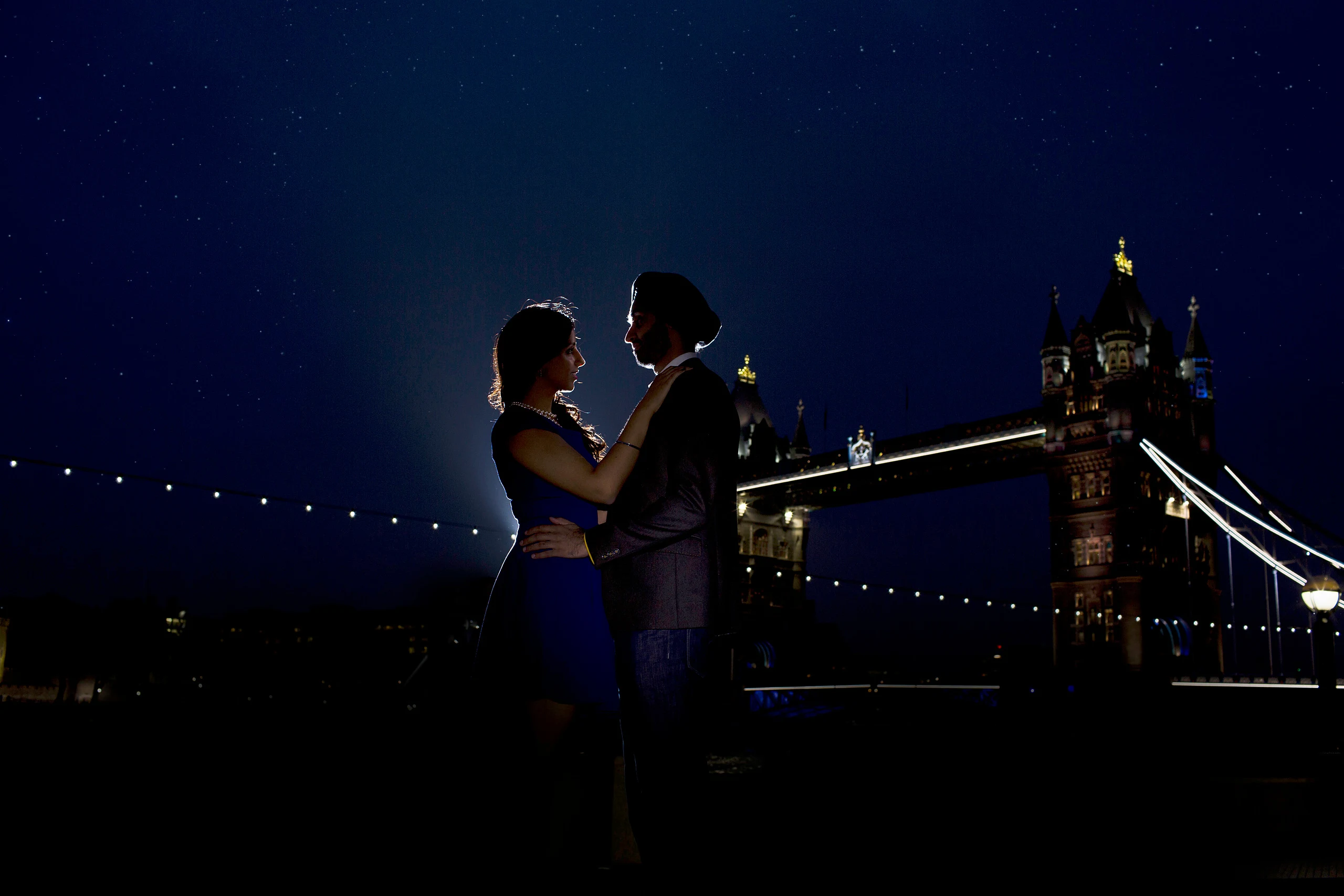 A couple embrace on the Thames riverside at night with Tower Bridge lit behind them under a starry blue sky.